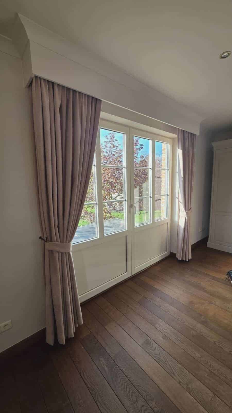 Vloerlange taupe gordijnen met sierlat en embrasse aan een tuindeur in een slaapkamer met donker parket – realisatie Aerts Interieur Lommel