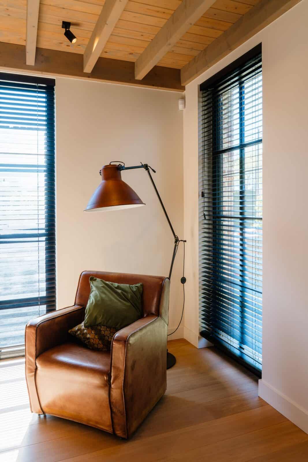 Donkere houten lamellen bij twee ramen met lederen fauteuil en industriële lamp - hobbykamer boven woning Geel