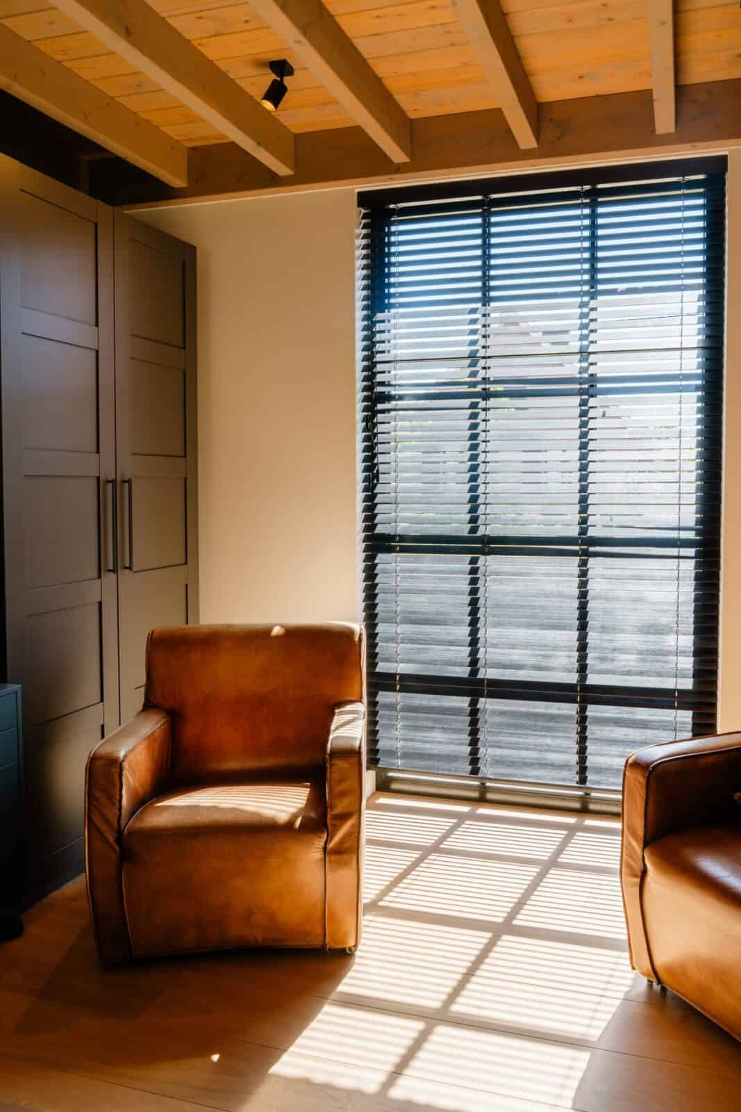 Donkere houten lamellen voor groot raam met lederen fauteuils en balkplafond - zithoek boven woning Geel