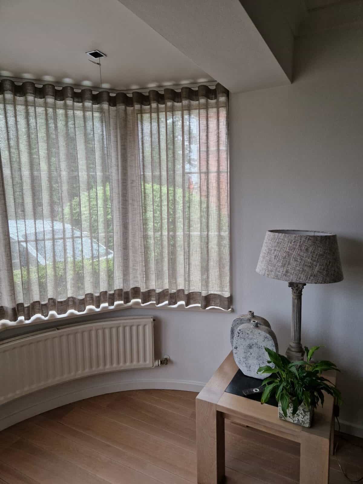 Taupe linnen wave gordijnen op gebogen rail in een ronde erker met parket en decoratieve lamp – realisatie Aerts Interieur Lommel