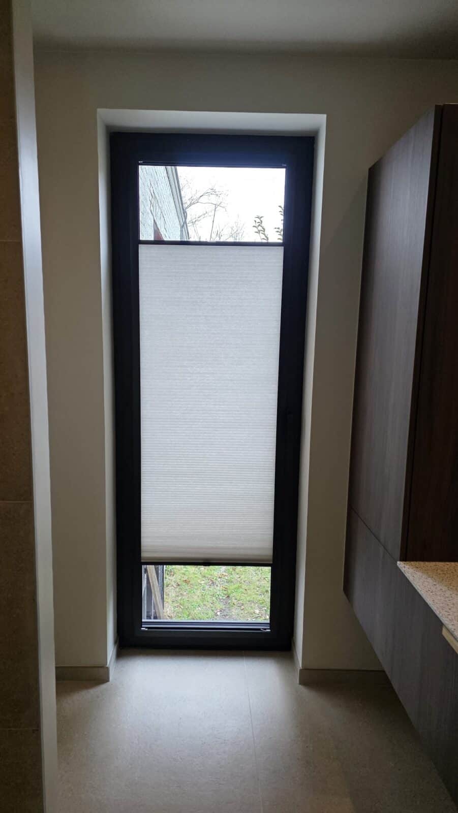 Duette plisségordijn halfopen op hoog smal raam – licht- en privacyregeling met zicht op tuin, geplaatst door Aerts Interieur Lommel
