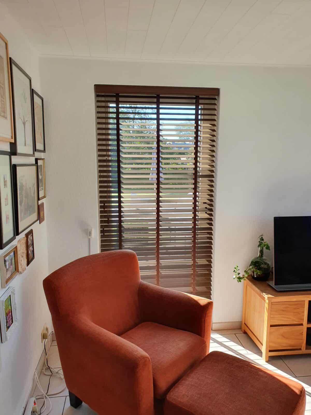 Donkerbruine houten jaloezie aan een hoog raam in een gezellige living met terracotta fauteuil en fotowand – realisatie Aerts Interieur Lommel